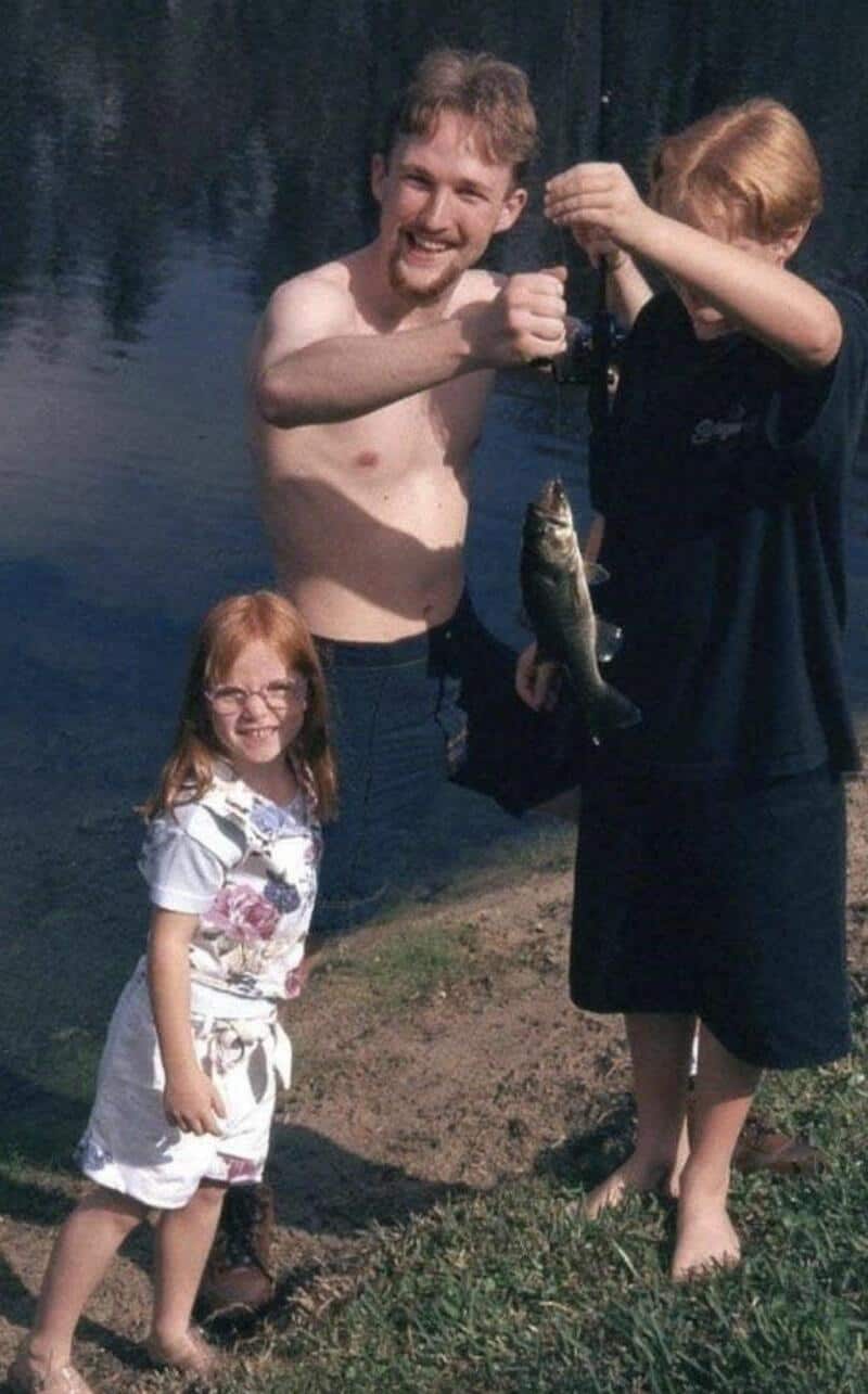 Two children and a man by a lake, one child holding a fish, all smiling.