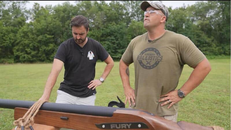 Two men standing outdoors near a wooden boat with a rope, one in black shirt, the other in green, in.