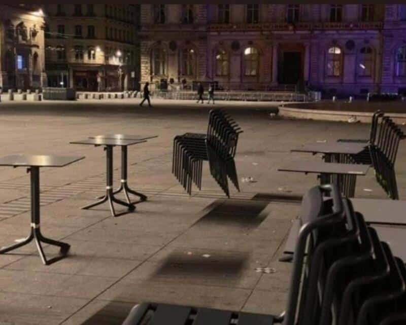 Empty outdoor seating area with stacked chairs casting shadows, city buildings in background.