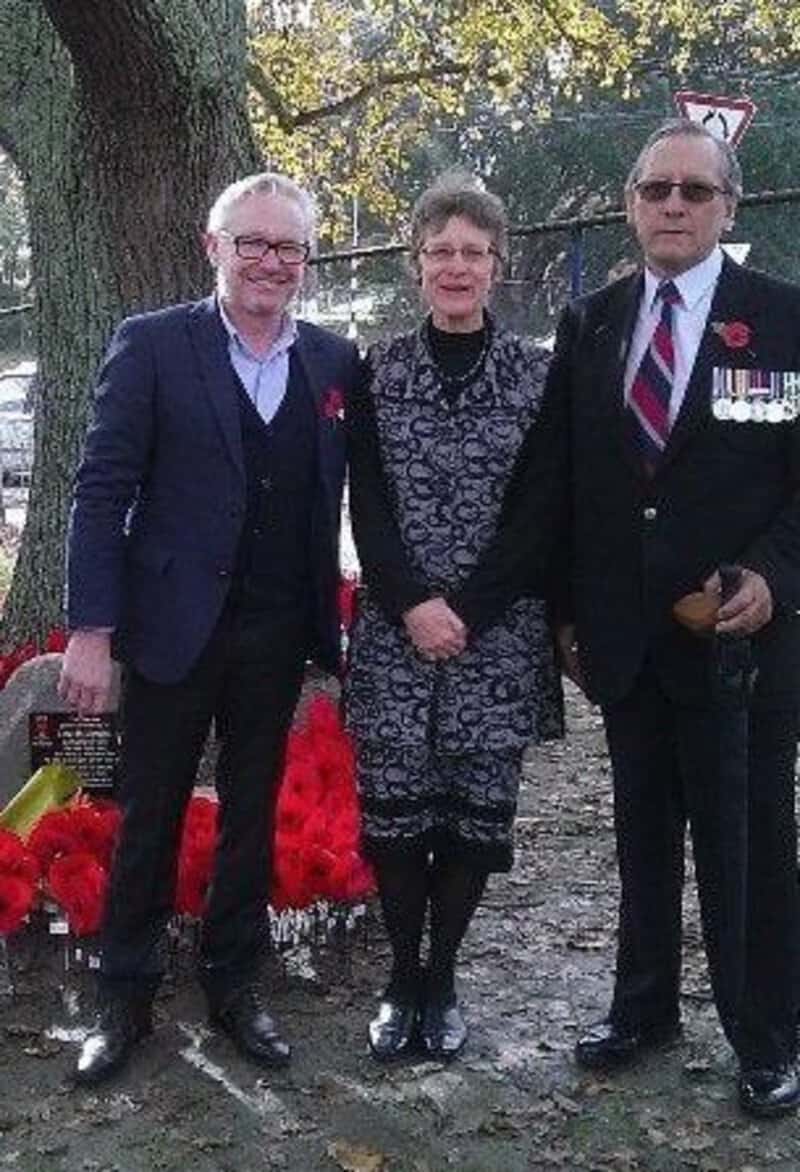 Three people standing outdoors, two men in suits and a woman in a dress, with a tree and a memorial.