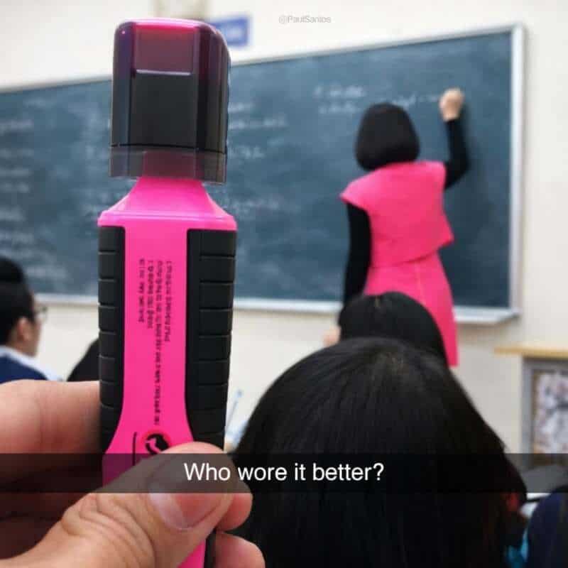 Pink highlighter held up in front of a classroom blackboard with a teacher writing.