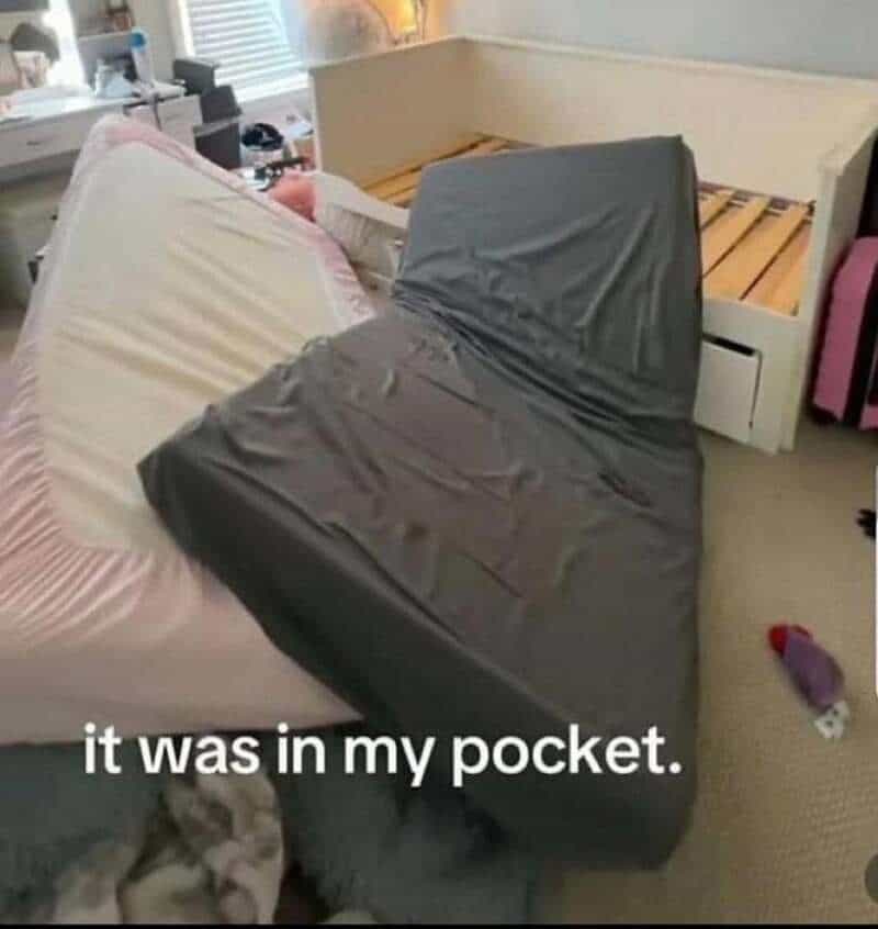 A black mattress is on the floor, leaning against a bed with a pink sheet, in a cluttered room.