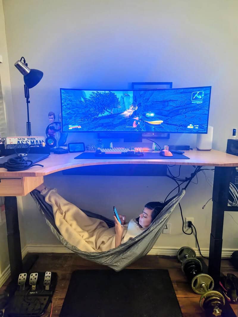 Person relaxing in hammock while playing video game on large monitor.