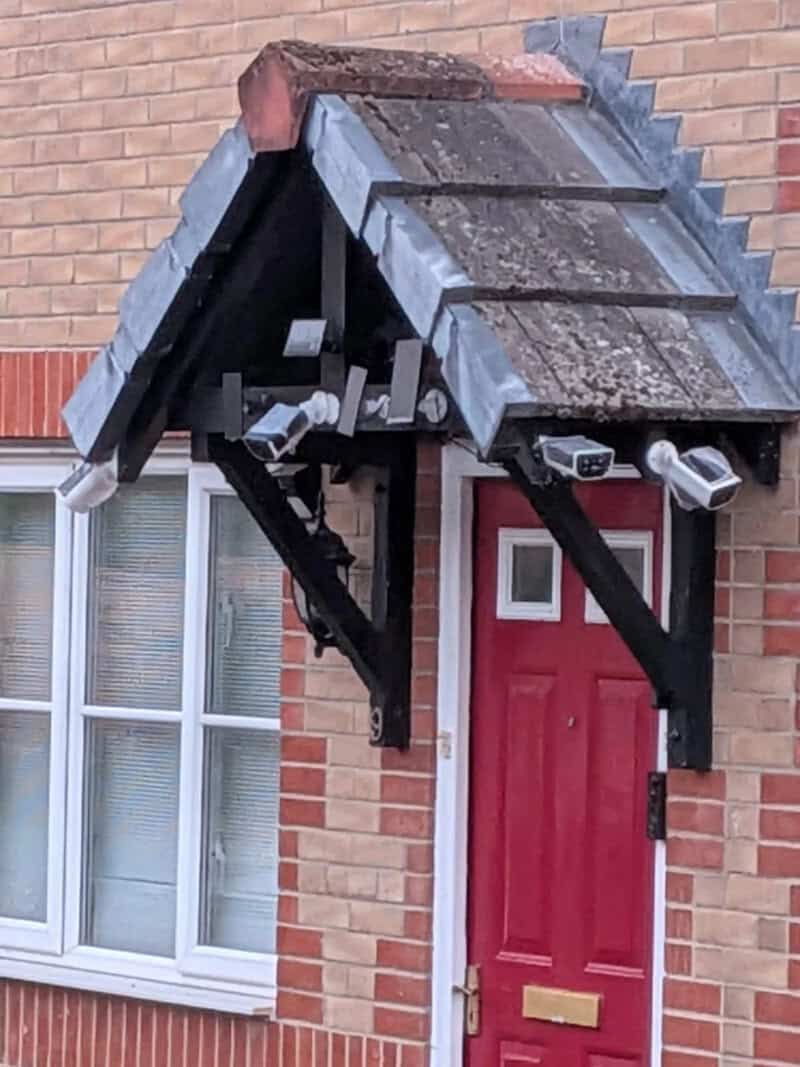 A small roof over a red door with a mailbox, cameras, and a pipe underneath.
