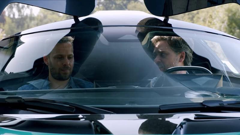 Two men sitting in a car, looking at each other through the windshield.