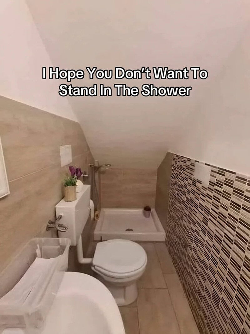 Small bathroom with a toilet, shower, and sloped ceiling, with text about shower standing.
