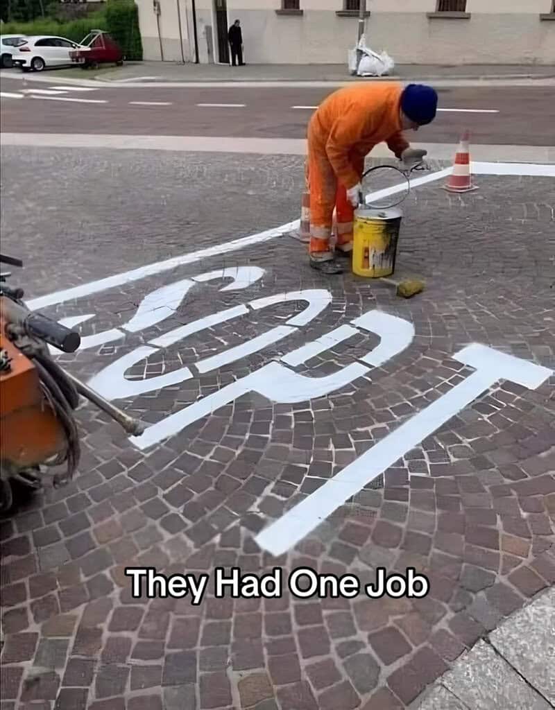 Worker painting a 'STOP' sign on cobblestone street with text 'They Had One Job'.