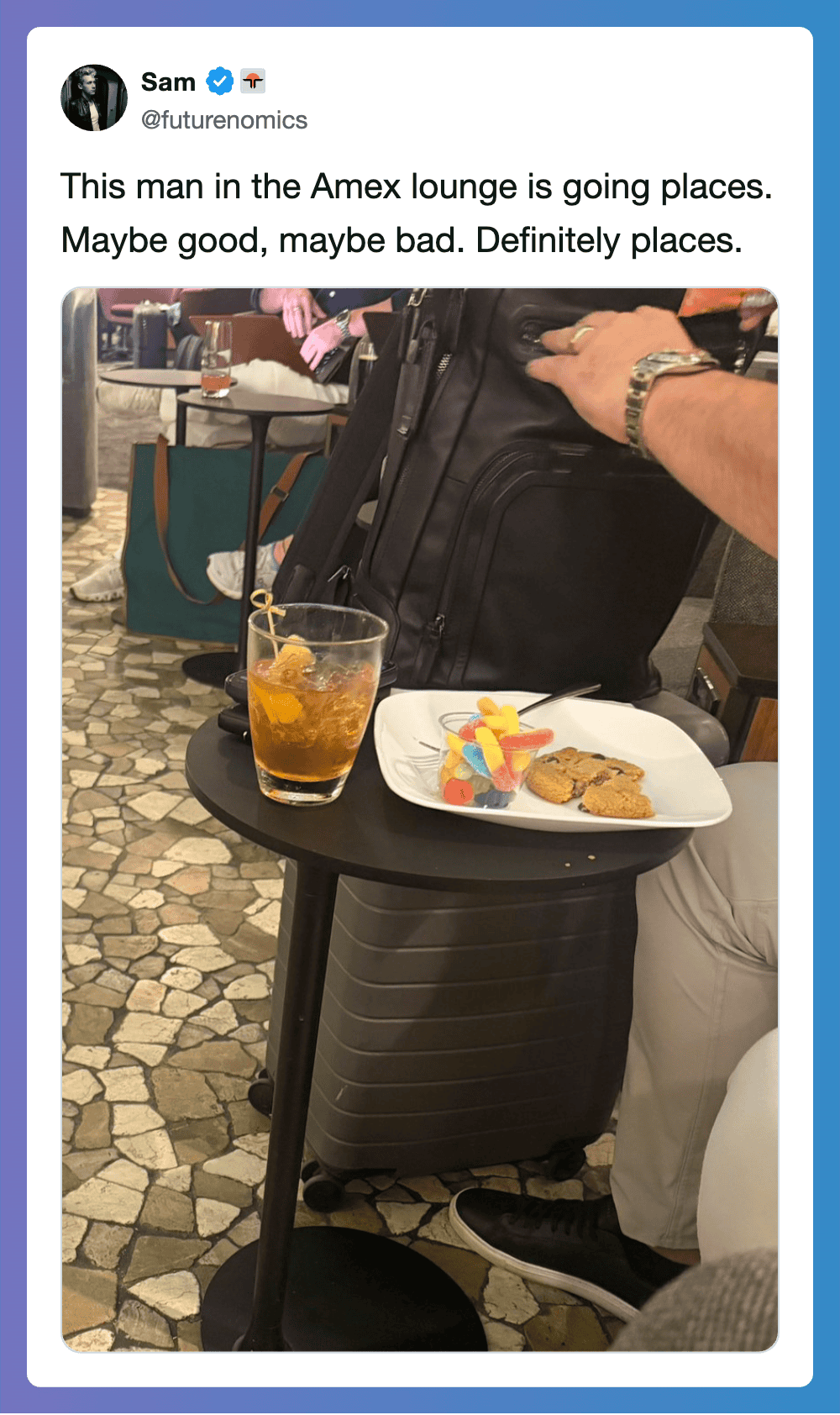 Man at Amex lounge with suitcase, drink, and snack on table.