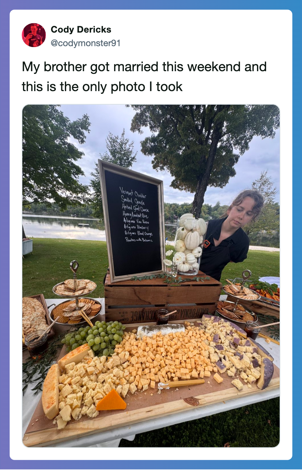 A woman at a lakeside outdoor event with cheese, grapes, and a blackboard menu.