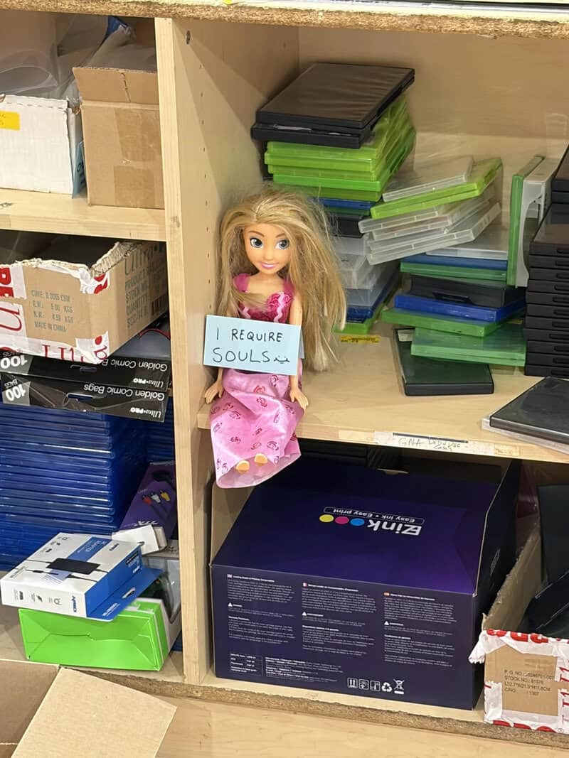 A doll holding a sign that says 'I REQUIRE SOULS' on a shelf with office supplies.