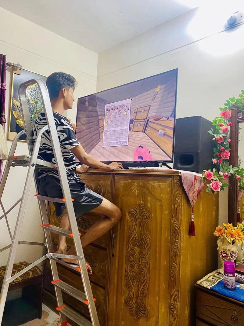 Young man playing a video game on a large TV, standing on a ladder in a living room.