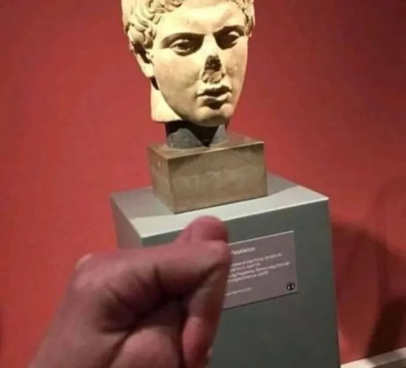 Ancient bust sculpture with a finger pointing at a plaque.