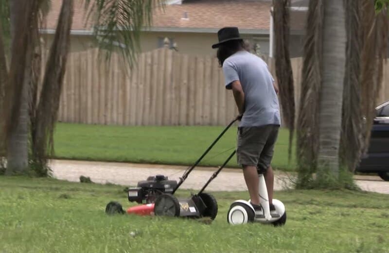 Person riding a hoverboard while mowing the lawn with a gas-powered mower.