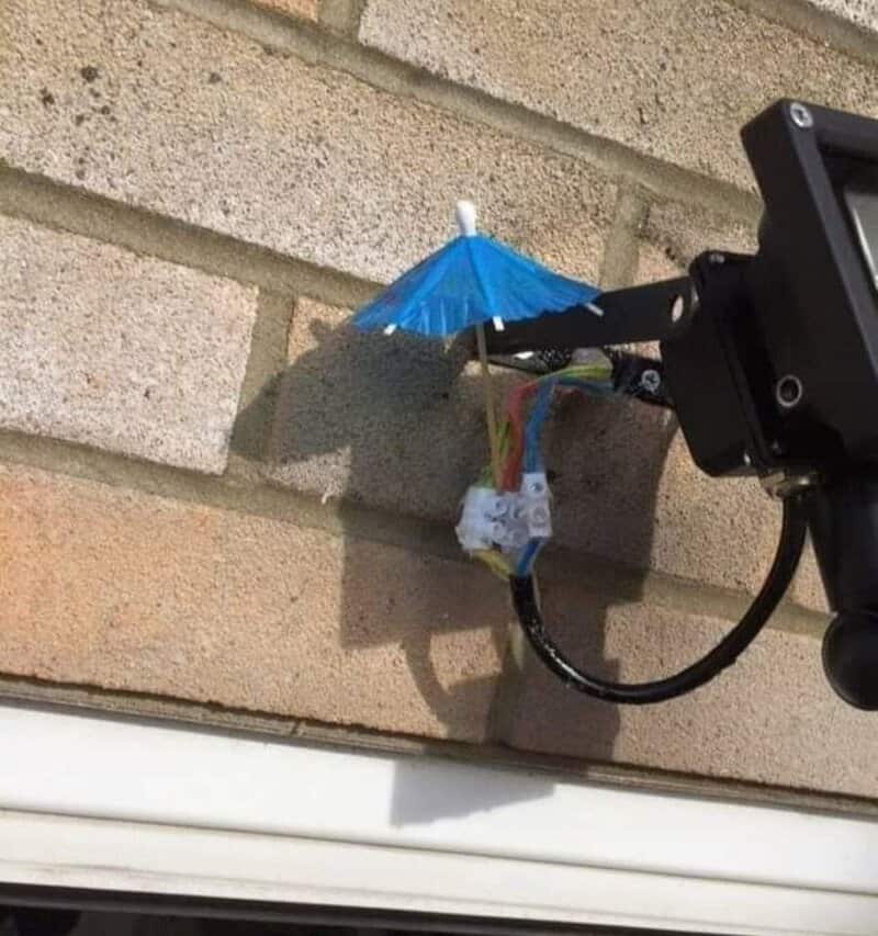 Miniature blue umbrella attached to a security camera on a brick wall.