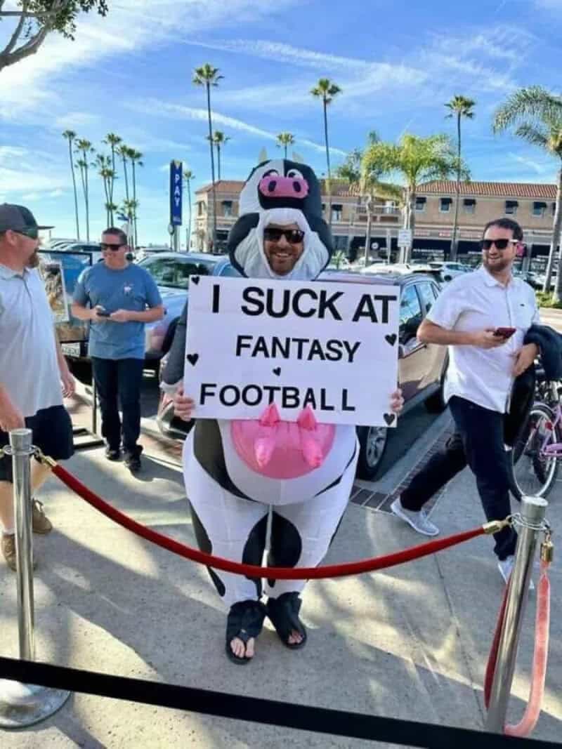 Person in cow costume holding sign that says "I SUCK AT FANTASY FOOTBALL".