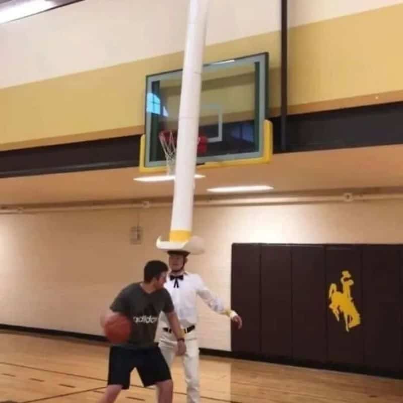 Person in a white suit with a hat balancing a long pole on their head in a gym.