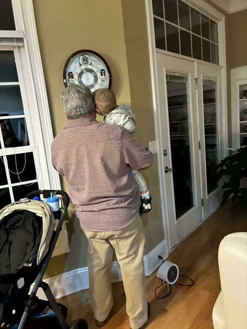 Grandfather holding a young child in a cozy home setting, looking at a clock.