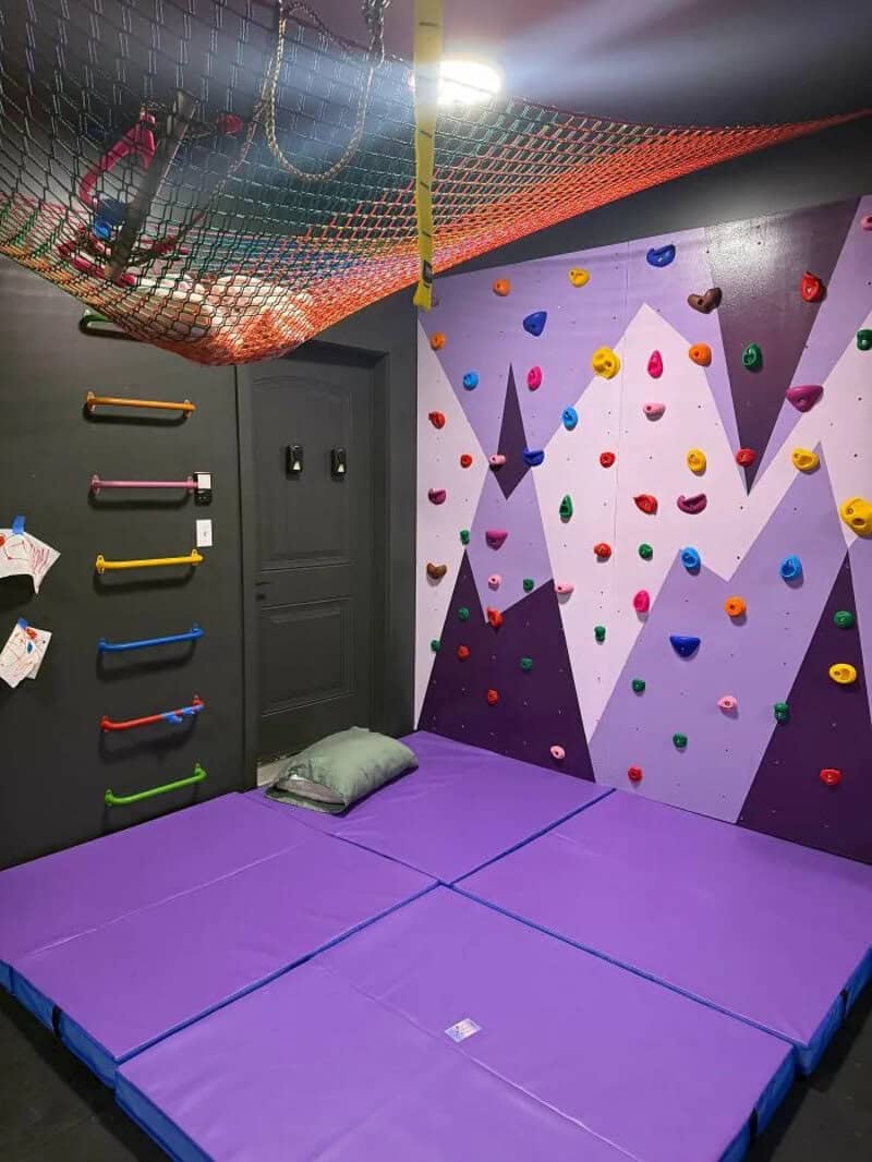 Indoor climbing gym with padded floor, colorful holds, and a safety net overhead.