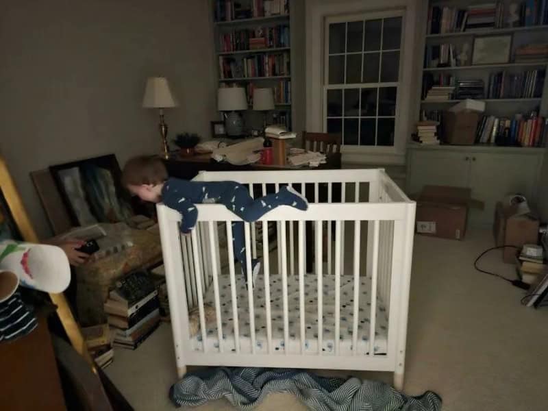 Child in pajamas climbing on a white crib in a cozy room.
