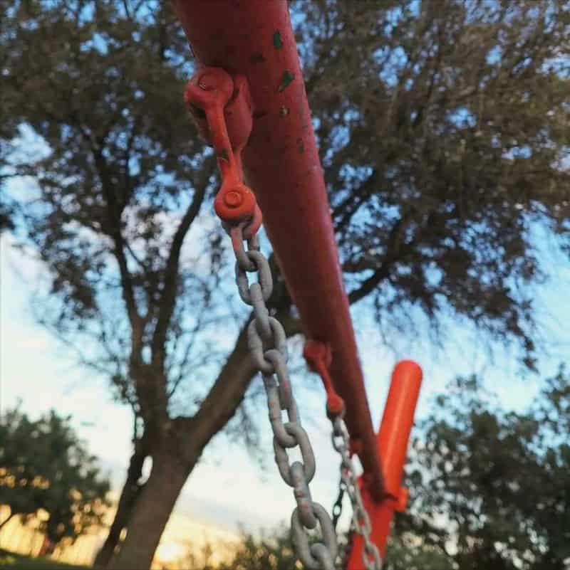 Close-up of a red swing set chain hanging from a red bar with trees in the background.