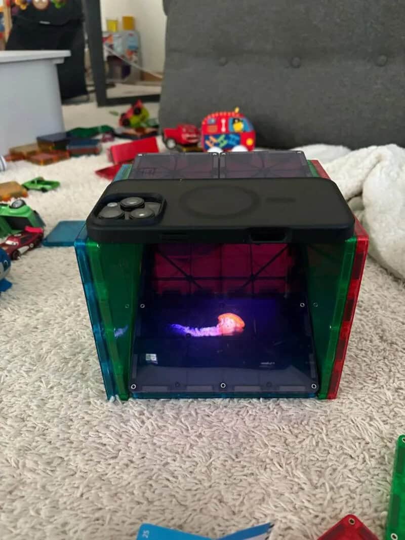 A small robot with a screen showing a colorful image, surrounded by toys on a carpet.