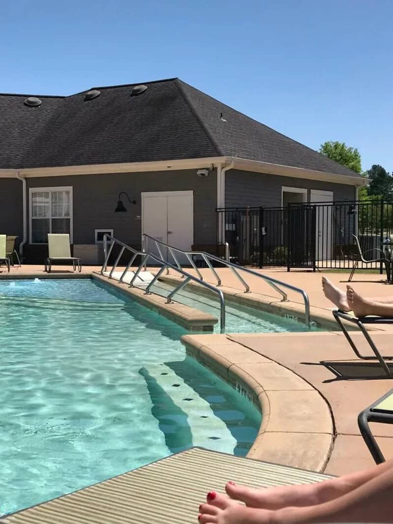 Pool with stairs, lounge chairs, and a building in the background under a clear blue sky.