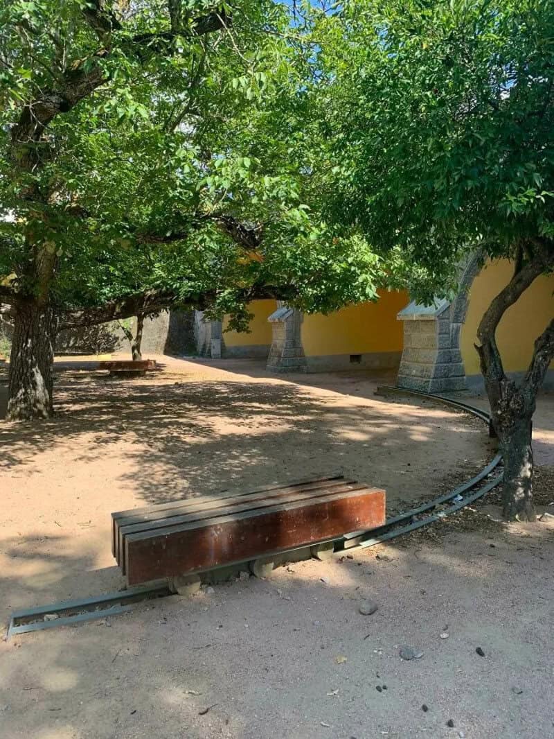 Broken park bench with curved metal support and green trees in background.