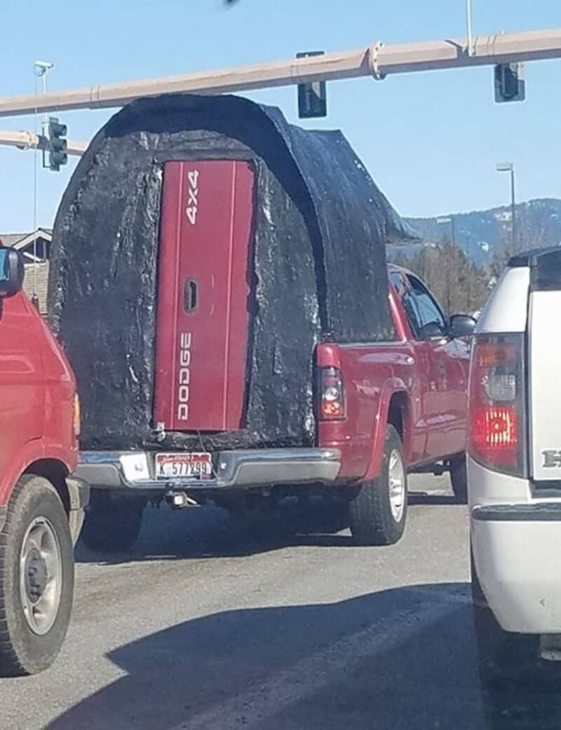 Red Dodge truck with a large rock on its bed, in traffic at an intersection.