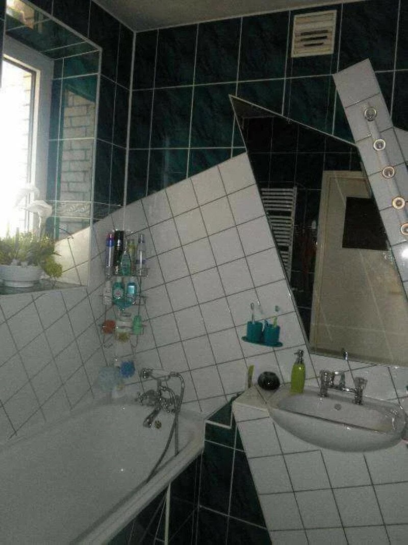 Bathroom with a tilted mirror and cluttered sink, tiles and window visible.