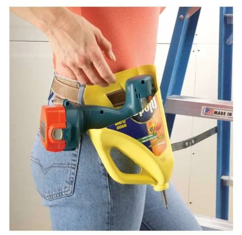 Person with a cordless drill in a yellow tool belt at a construction site.