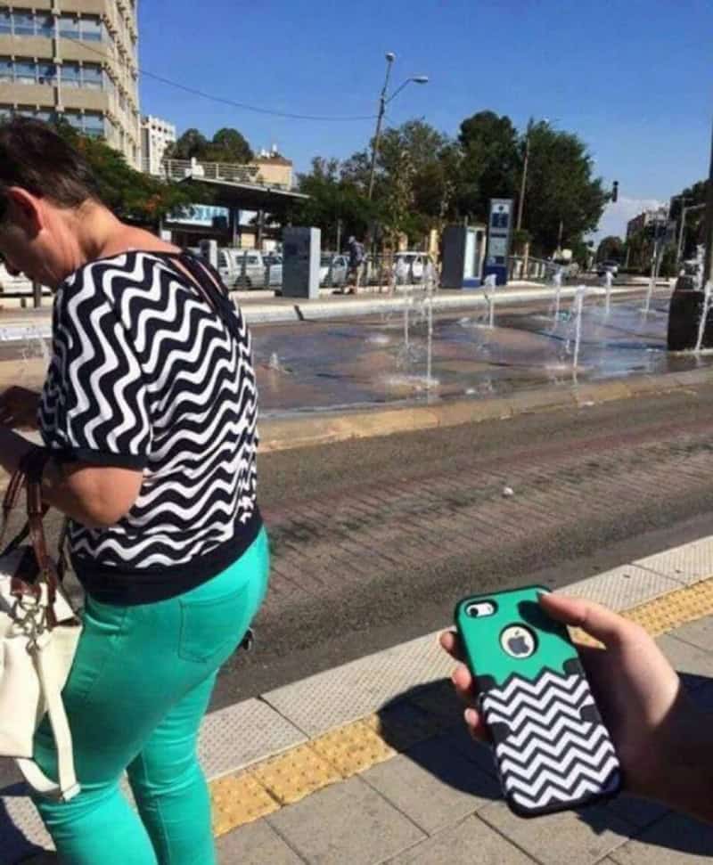 Person taking a photo of a woman near a fountain on a city street.
