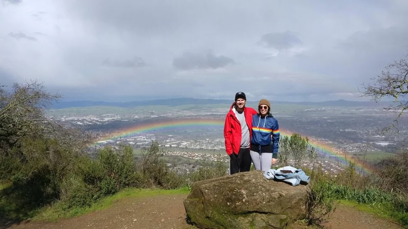 Two people stand on a hill with a rainbow in the background.