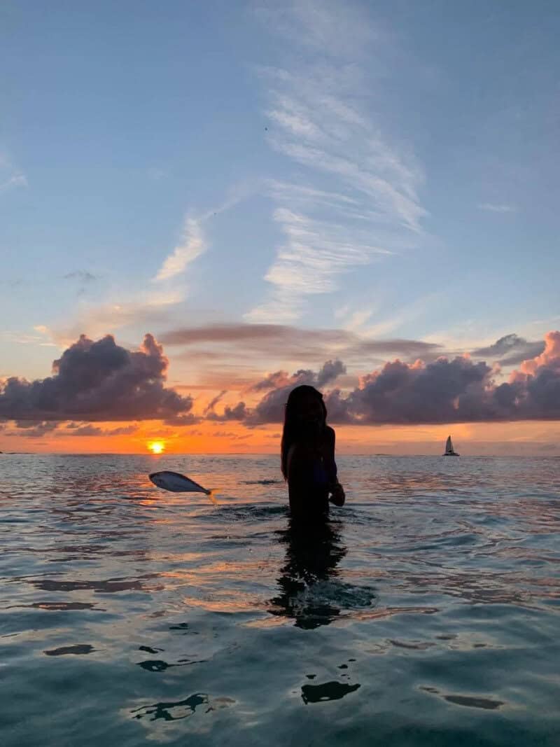 Silhouette of a woman in water at sunset with a paddleboard and sailboat in the background.