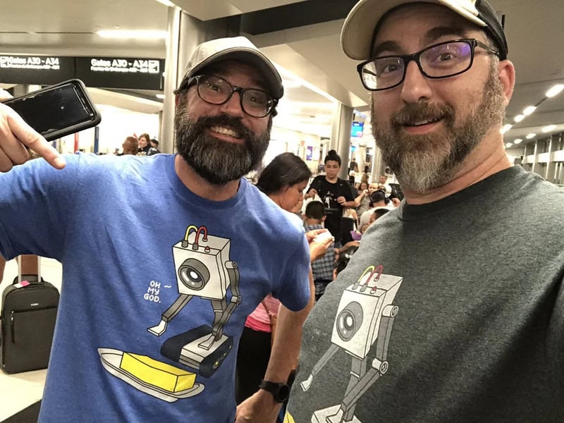 Two men in glasses and caps smiling at the airport, wearing T-shirts with a washing machine cartoon.