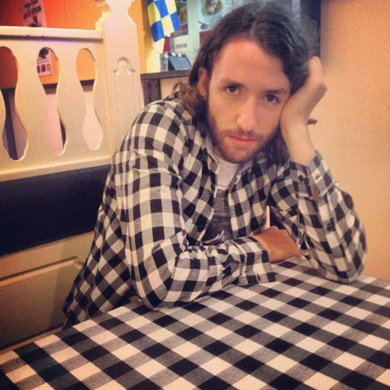 Man with long hair and checkered shirt sitting at a table in a restaurant.