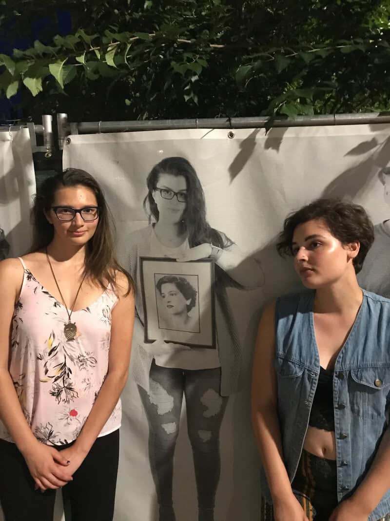 Two women stand in front of a poster of a woman holding a framed photo.