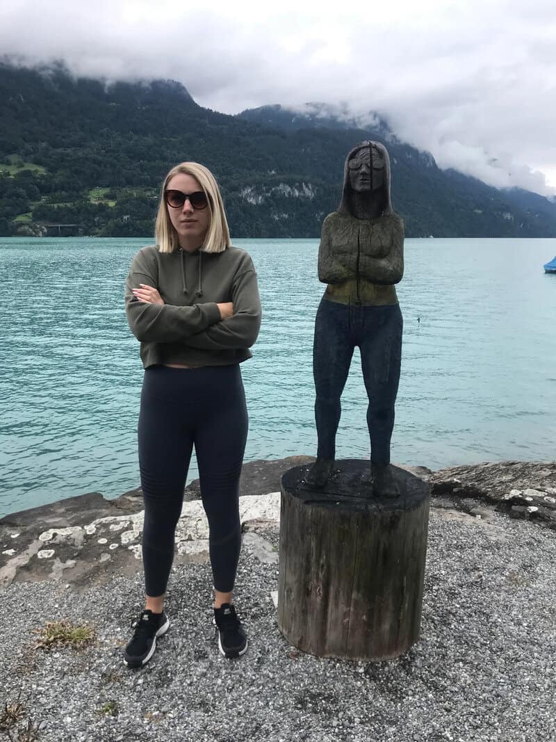 Woman with sunglasses standing next to a wooden sculpture of a woman by a lake.
