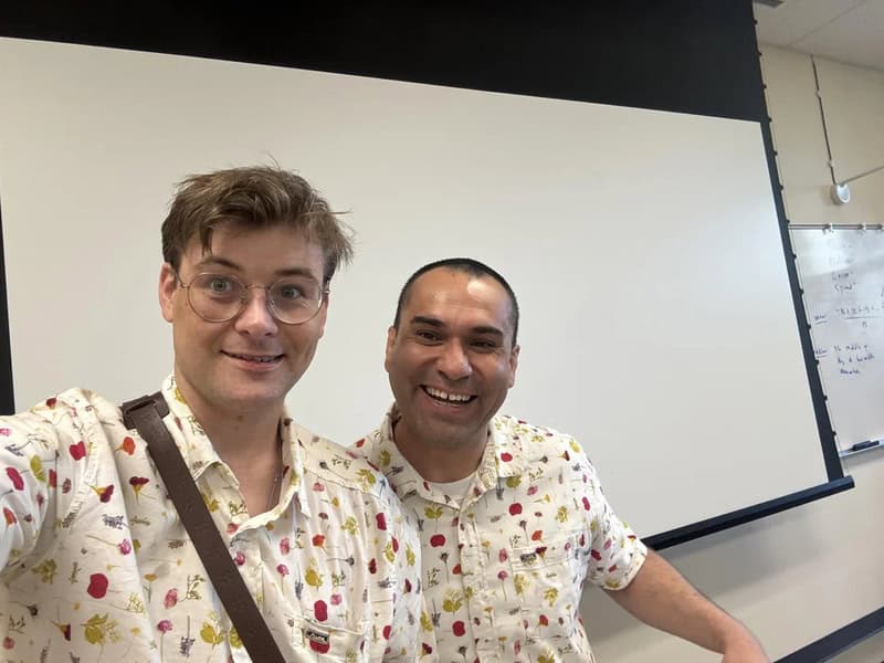 Two men smiling for a selfie in a classroom with a whiteboard behind them.