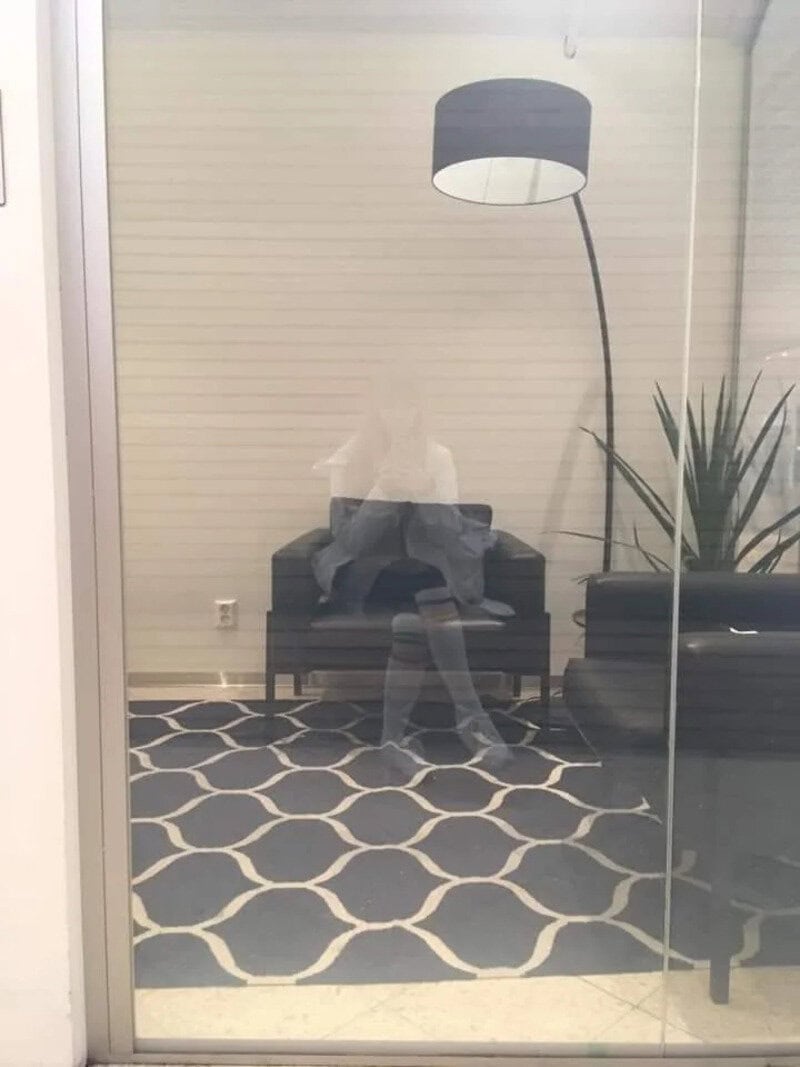 Person sitting on a black chair reflected in glass door with a lamp and plant nearby.