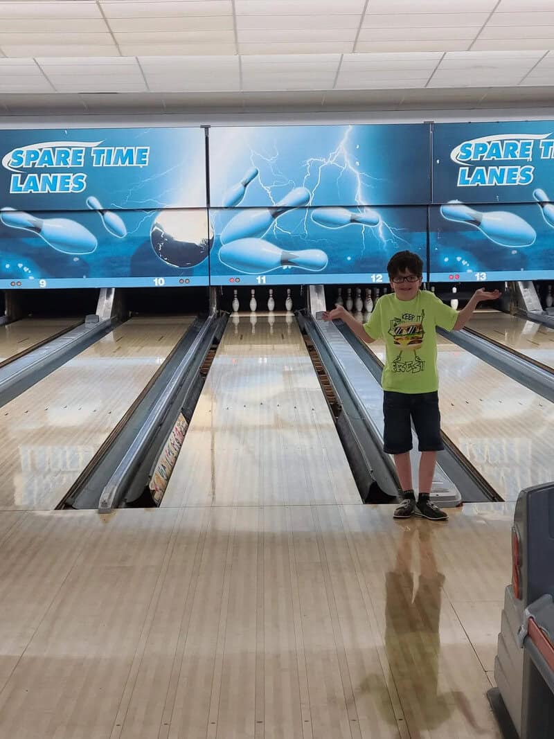 Child standing in bowling alley with lightning-themed backdrop and 'Spare Time Lanes' signs.