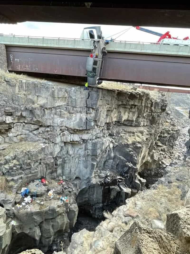Car hanging from bridge over a deep rocky chasm with trash below.