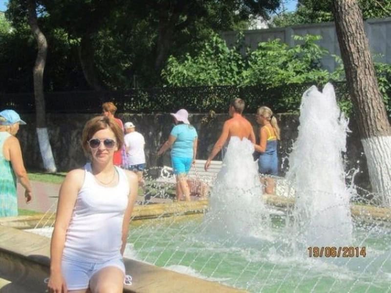 Woman in white tank top and sunglasses near water fountain with people in background.