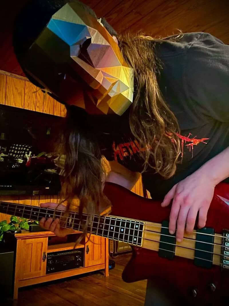 Person with a colorful geometric mask playing a bass guitar in a room.