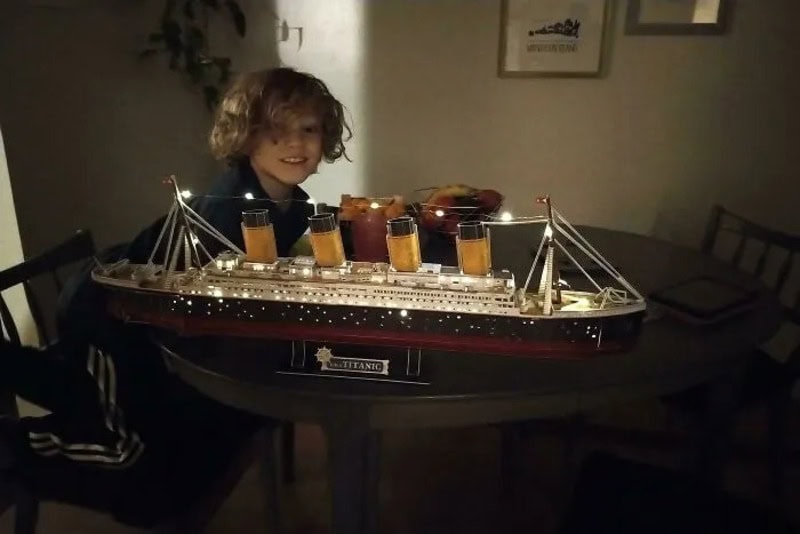 Child with curly hair smiling at a model of the Titanic on a table.