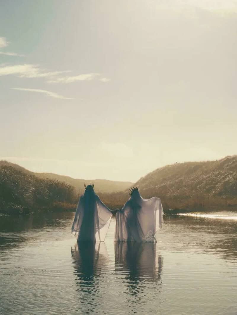 Two figures in flowing garments stand in a river at sunset, holding hands.