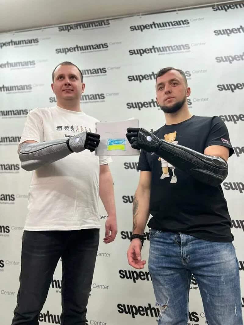 Two men with prosthetic arms holding a small flag at Superhumans Center.