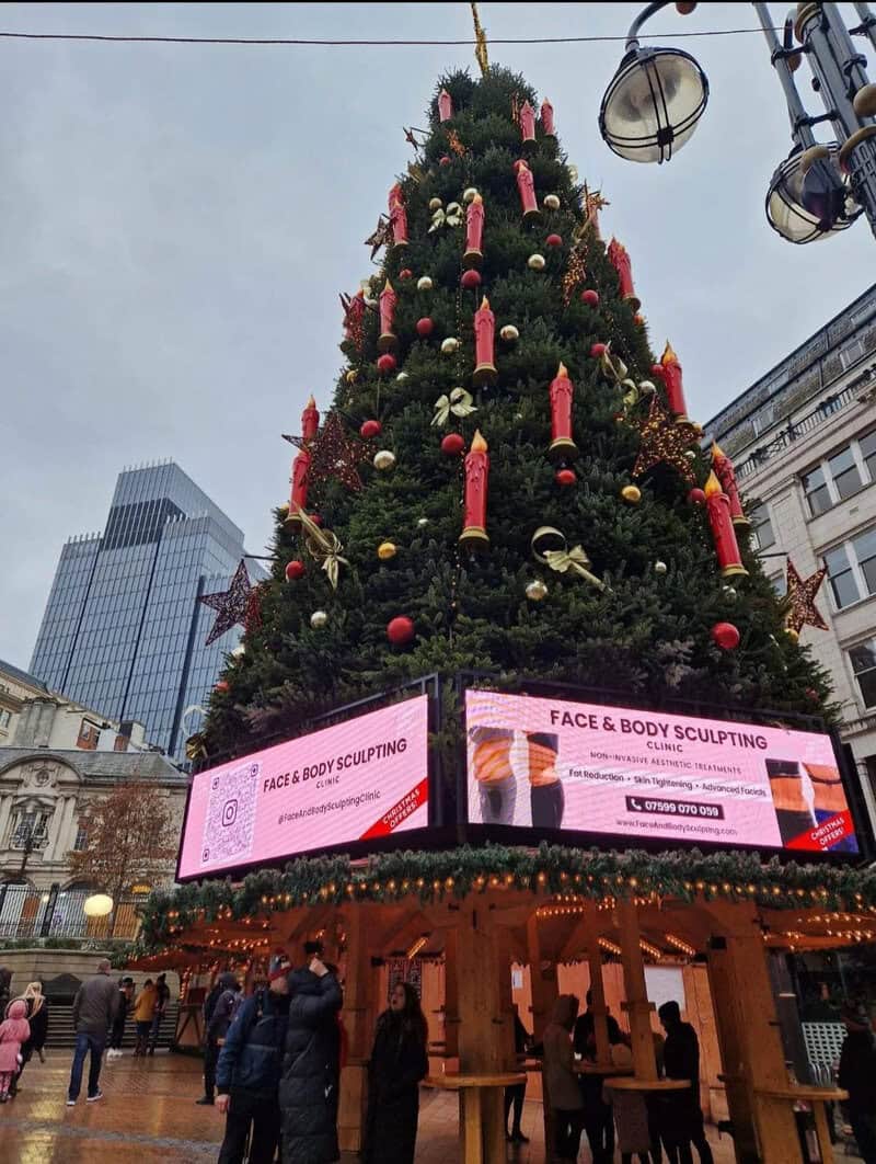 Large decorated Christmas tree in an urban square with digital advertising screens at the base.