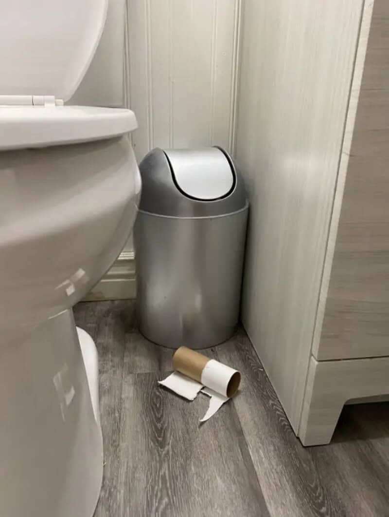 Trash can with toilet paper roll and discarded paper on floor next to toilet.