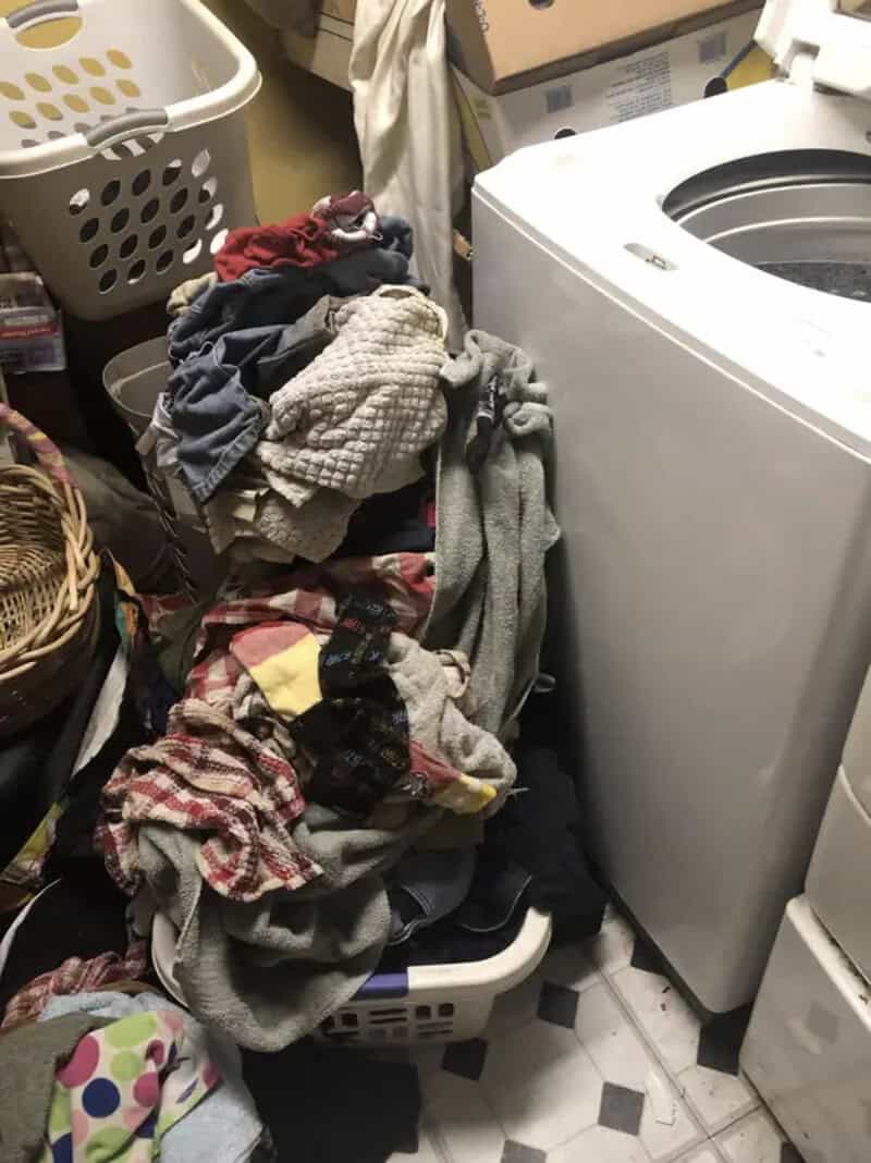 Clothes and laundry piled next to a washing machine in a cluttered laundry room.