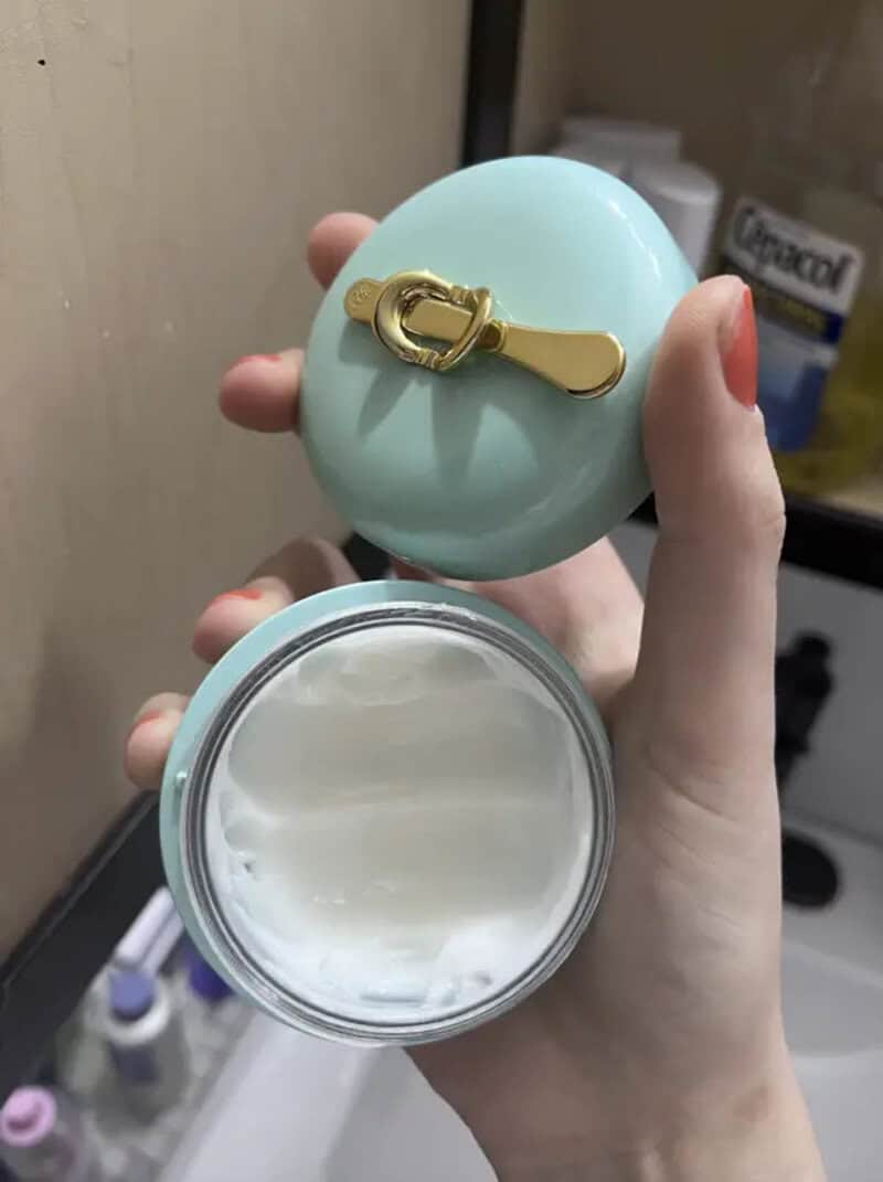 A hand holding a round container with a light blue lid and a gold zipper pull.